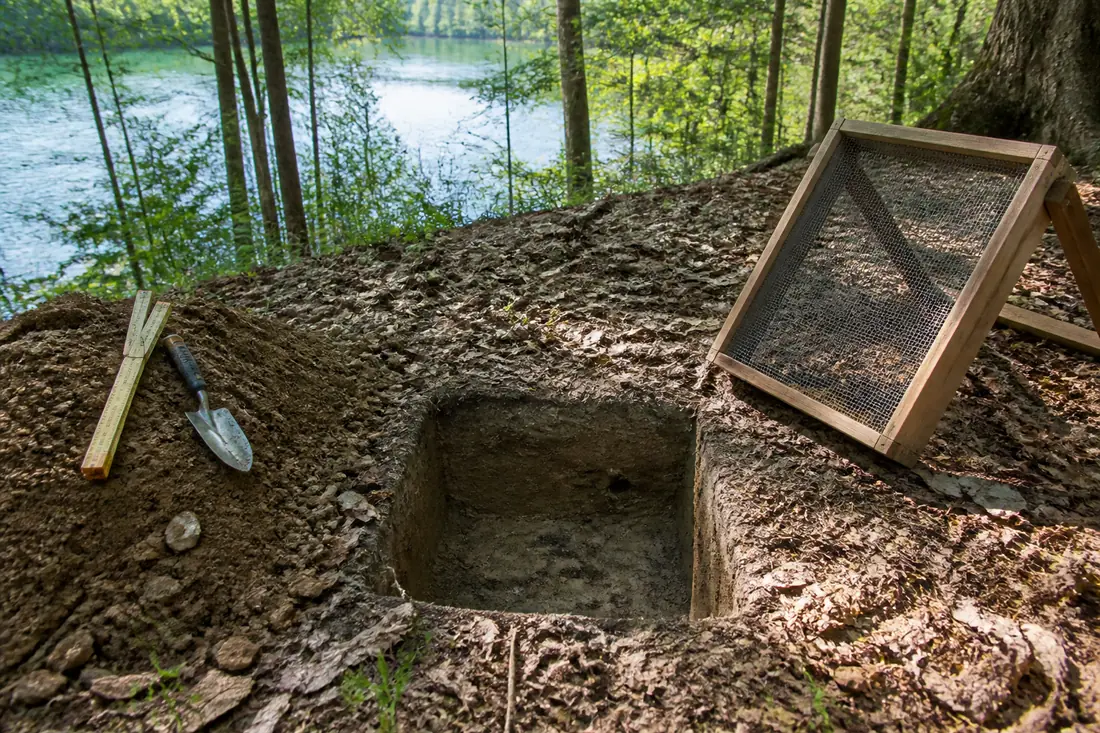 A small square test excavation on a wooded bank with a screen and trowel beside it