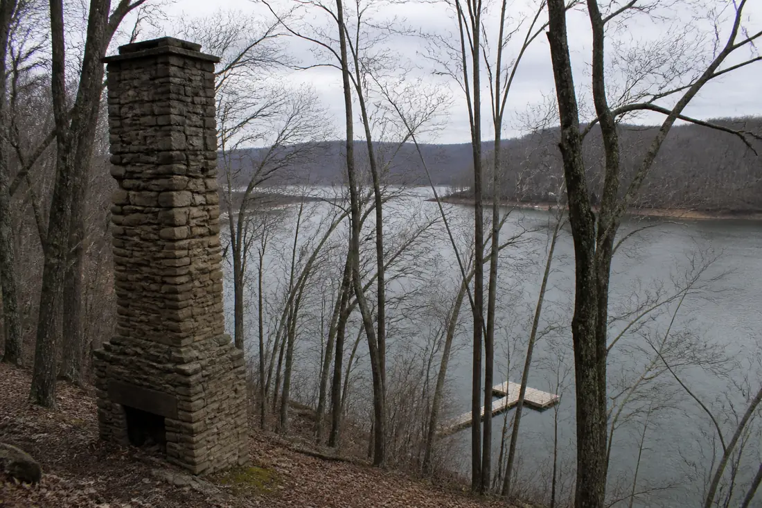 A stone chimney standing alone on a wooded reservoir bank above a dock